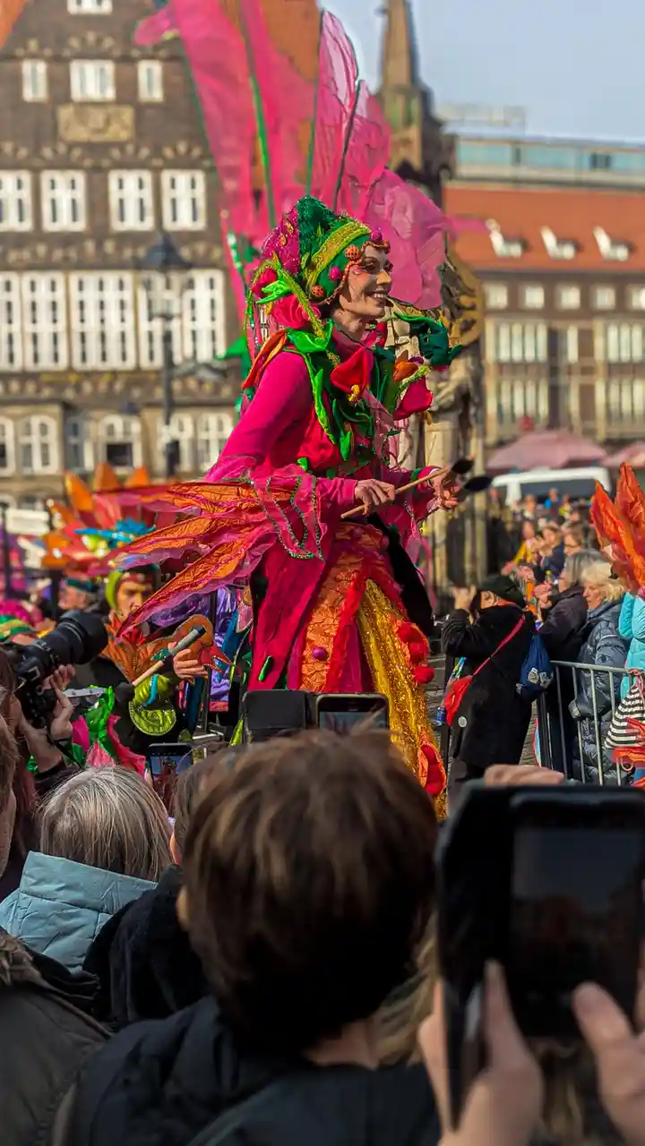 A person in a vibrant, elaborate costume with pink, green, and orange fabric and large feather-like wings smiles while…