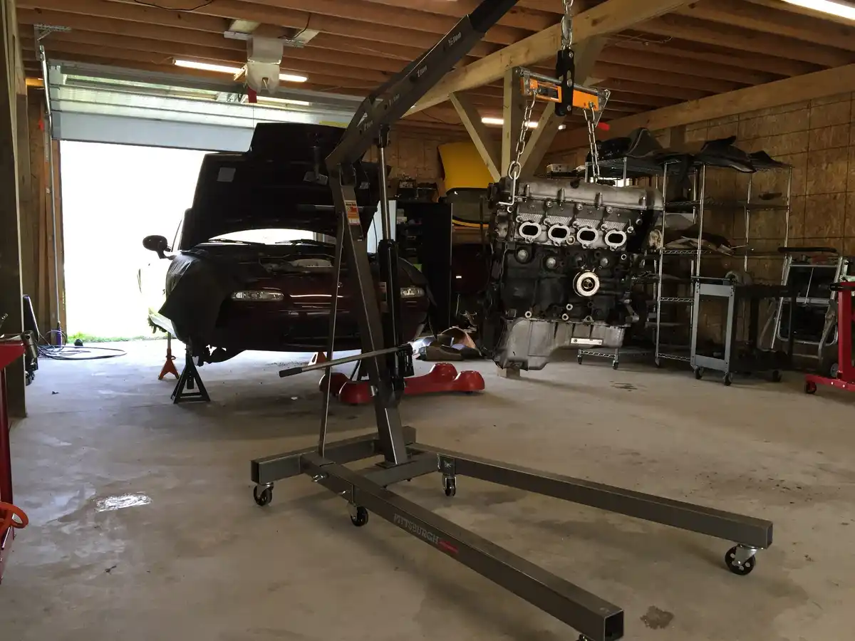 A garage scene shows a car with its hood open and a large engine suspended by chains from a crane. The engine is being…