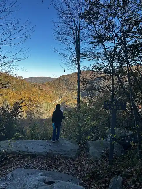 A person in a black hoodie and blue jeans stands on a large rock overlooking a scenic autumn forest valley under a clear…