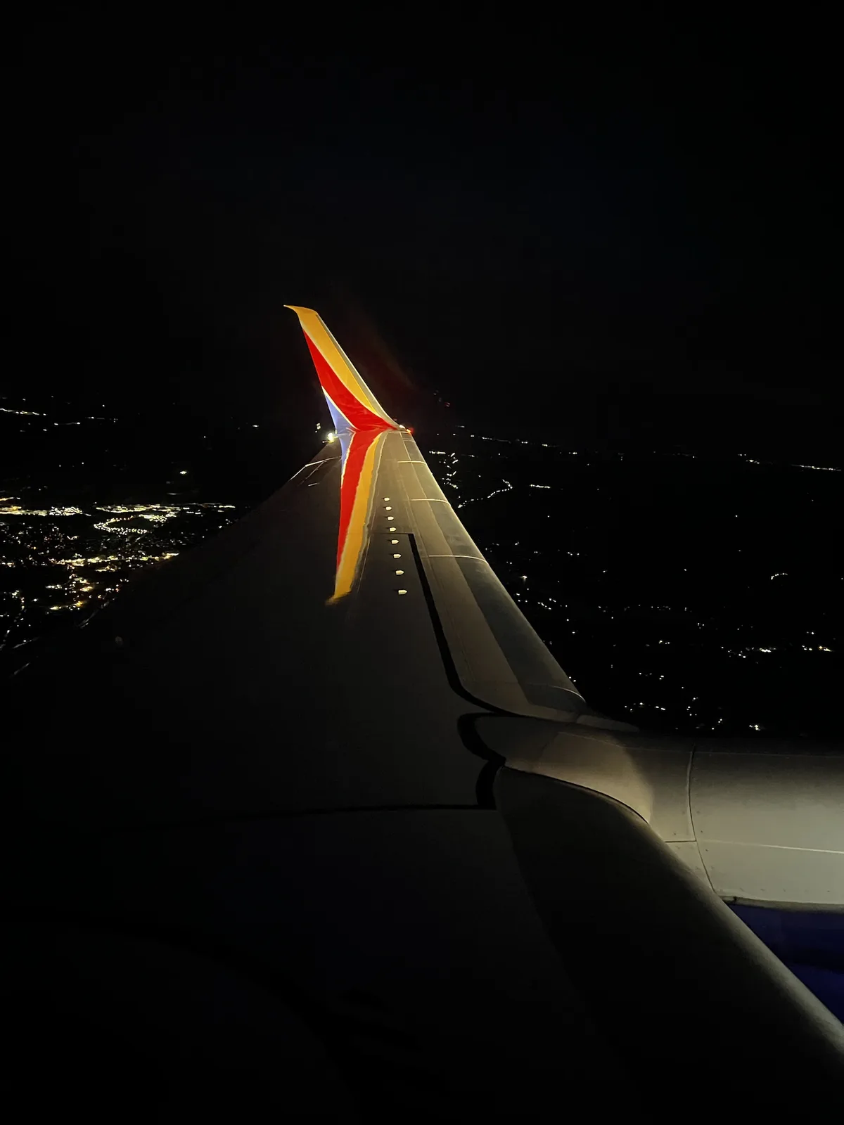 A nighttime photo taken from an airplane window showing the wing and tail of a Southwest Airlines plane, with city lights…
