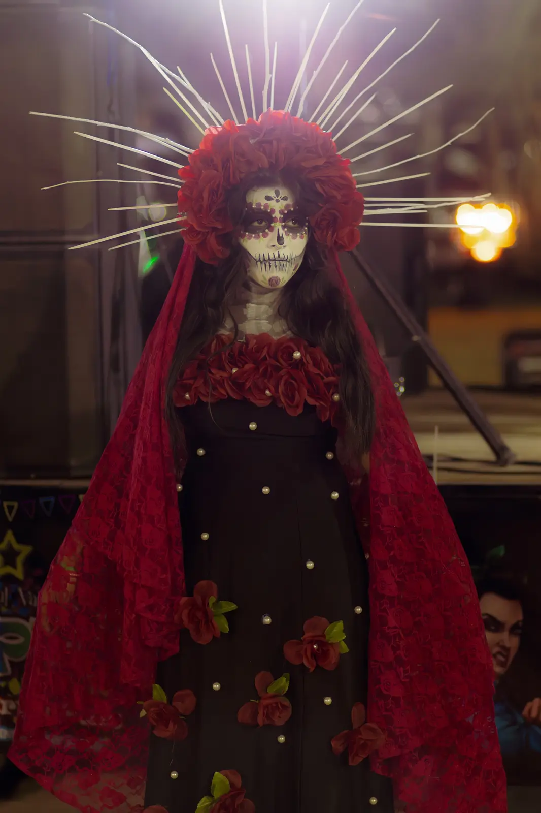 A person dressed as a Day of the Dead figure with skull makeup, a red floral headdress with white spikes, and a black dress…