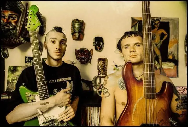Two men, one holding a green electric guitar and the other a wooden bass guitar, stand in a room decorated with masks and…