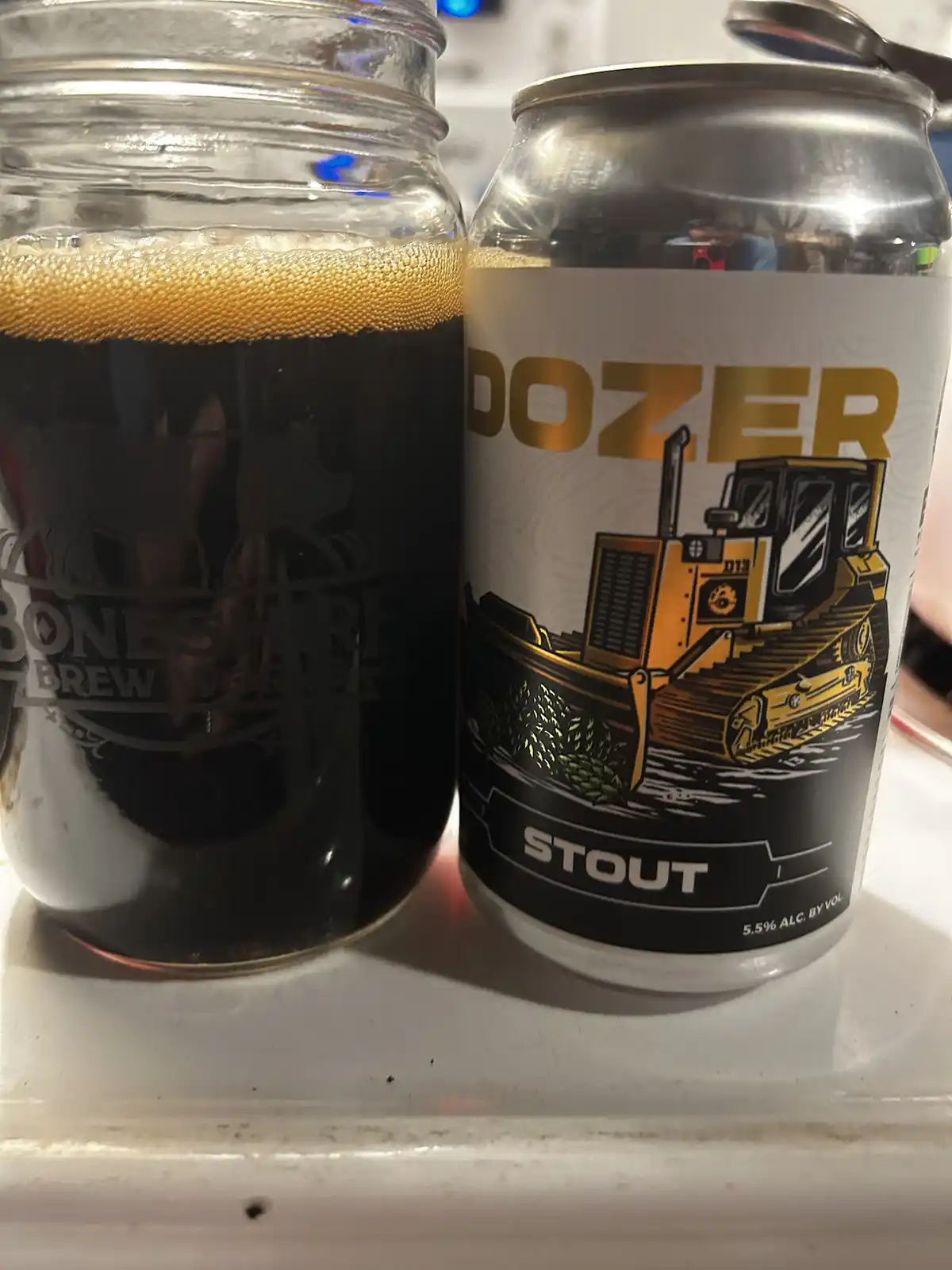 A can of 'Dozer Stout' beer, featuring a yellow bulldozer illustration, sits next to a mason jar filled with dark beer…