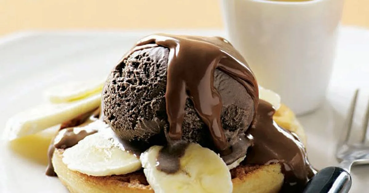 A close-up of a dessert featuring a scoop of chocolate ice cream melting on top of a pancake or waffle, generously drizzled…