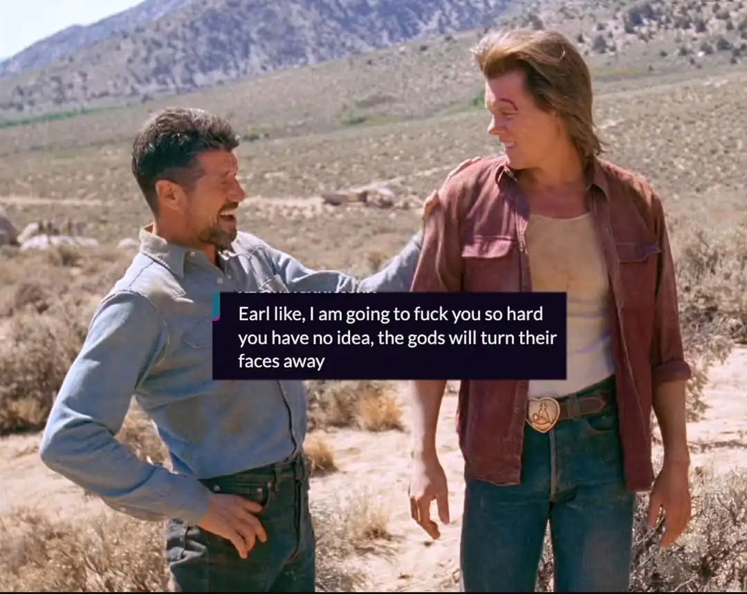 Two men stand in a desert landscape, one in a blue shirt with a hand on the other's shoulder, who has a bruise on his face…