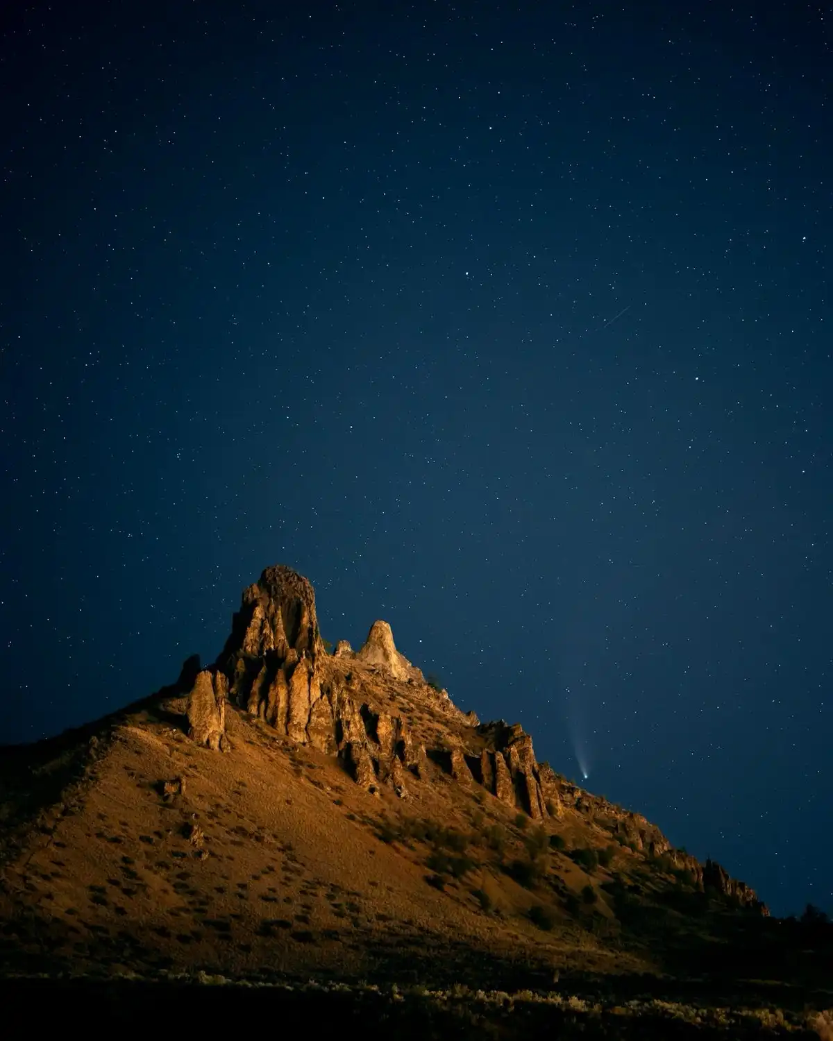 A night photograph shows a rugged, rocky mountain peak under a star-filled dark blue sky. A bright, glowing comet with a…
