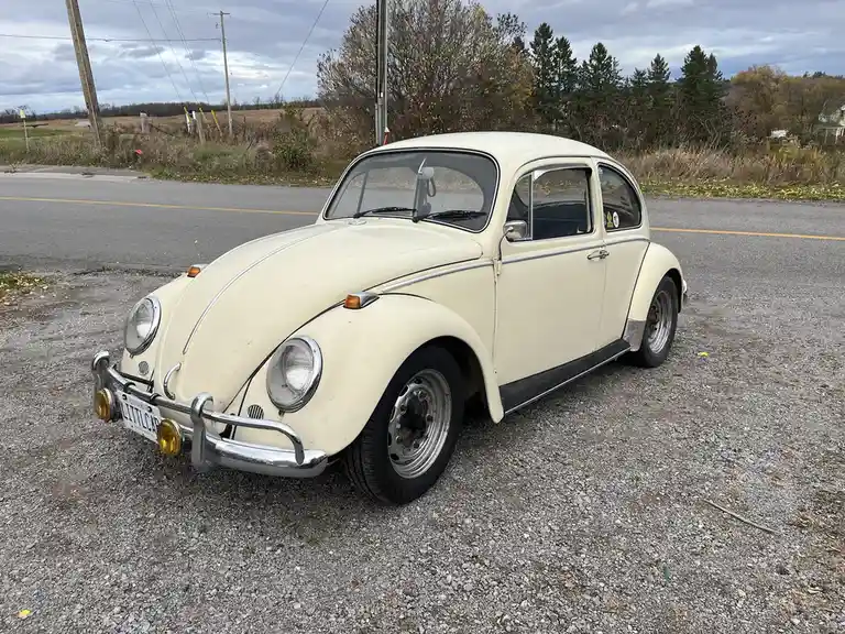 A cream-colored vintage Volkswagen Beetle is parked on a gravel shoulder beside a road, with a cloudy sky and trees in the…