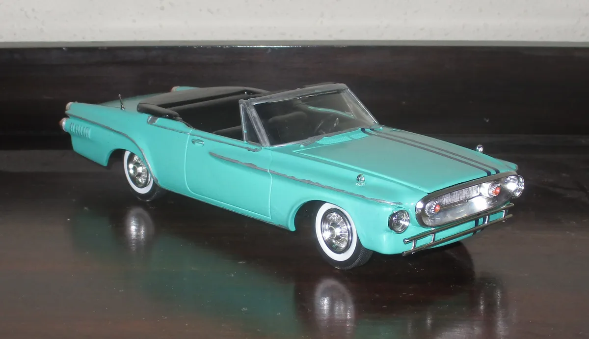A turquoise vintage convertible toy car with black interior and white-wall tires sits on a dark reflective surface. The car…