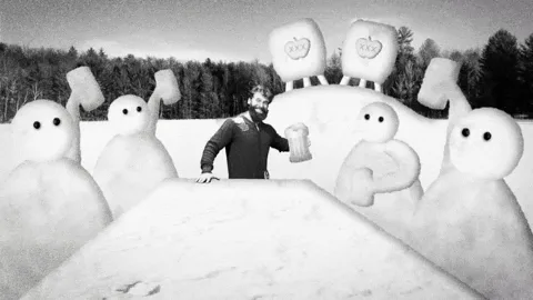 A black-and-white image shows a man in a dark jacket standing behind a snowbank, surrounded by cartoonish snowmen. In the…