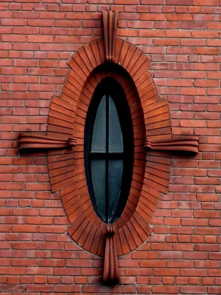 a close-up photograph of an ornate, vertically elongated oval window set in a red brick wall, framed by decorative brickwork…