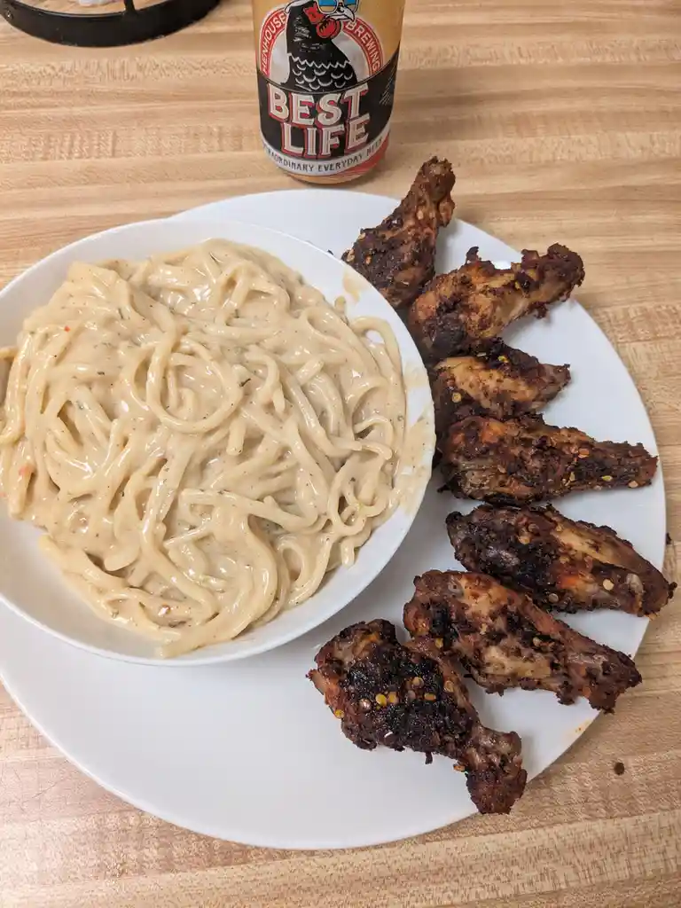 A plate of creamy pasta and crispy chicken wings sits on a wooden table next to a can of 'Best Life' beer, with the text…