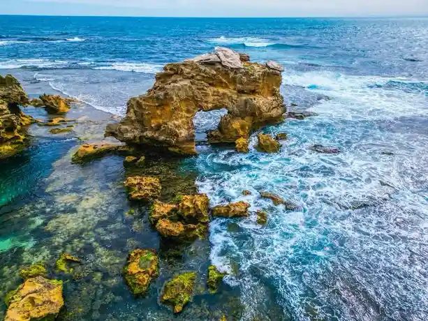 A scenic photo of a natural rock arch formation in the ocean, with turquoise water, crashing waves, and yellowish-brown…