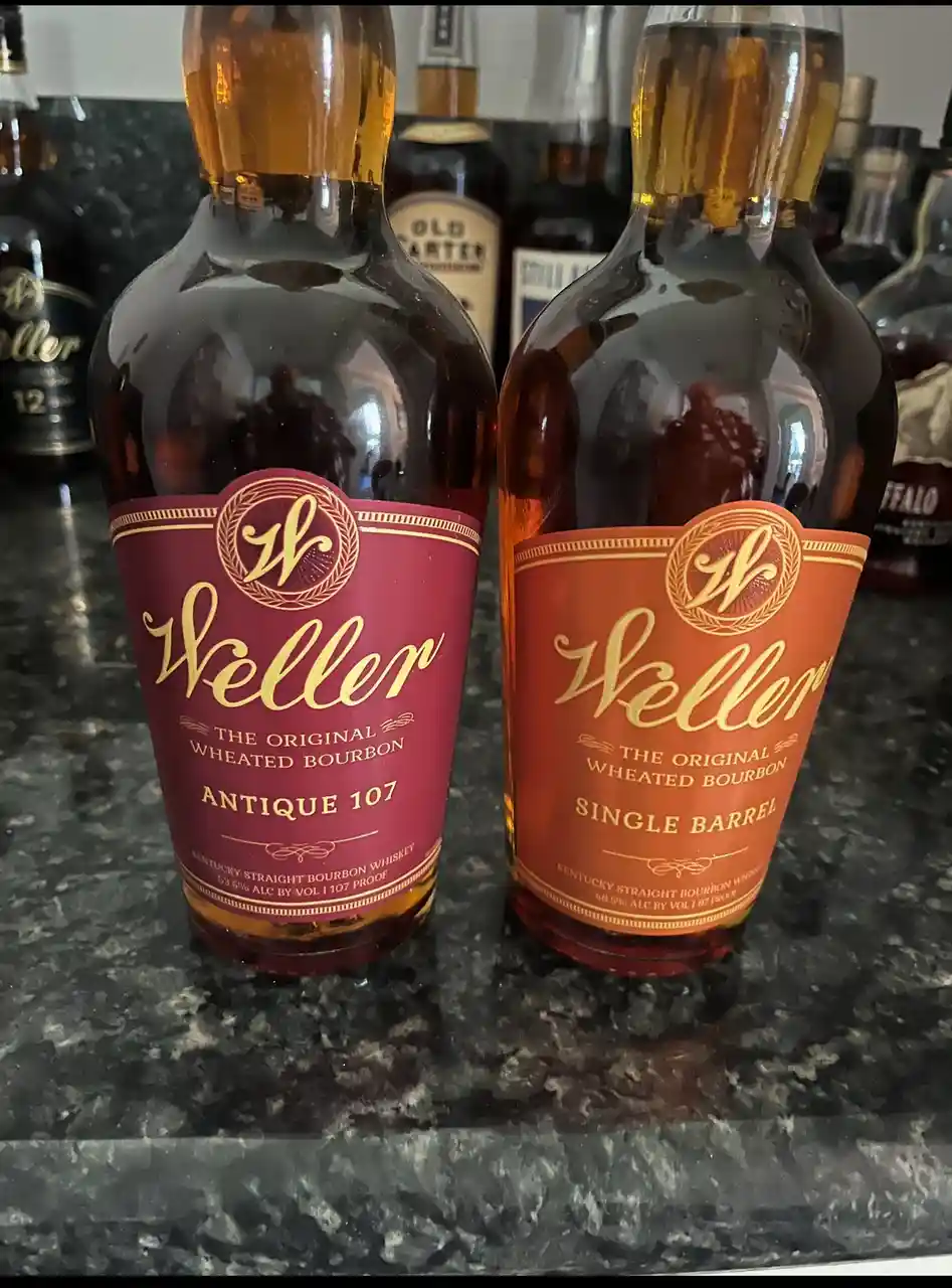 Two bottles of Weller bourbon whiskey sit side-by-side on a dark countertop, one with a red label labeled 'Antique 107' and…