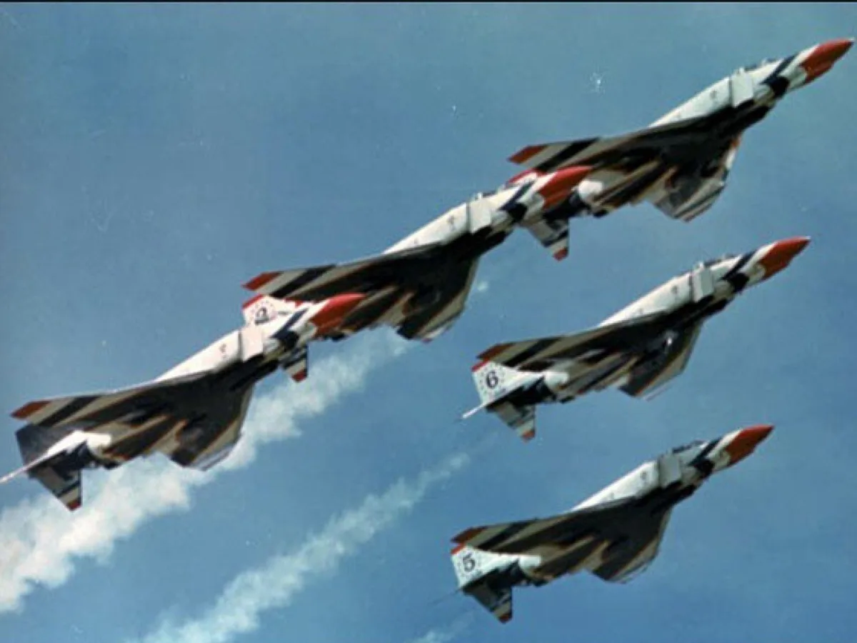Three fighter jets with red, white, and black camouflage patterns fly in formation against a blue sky with wispy clouds. The…