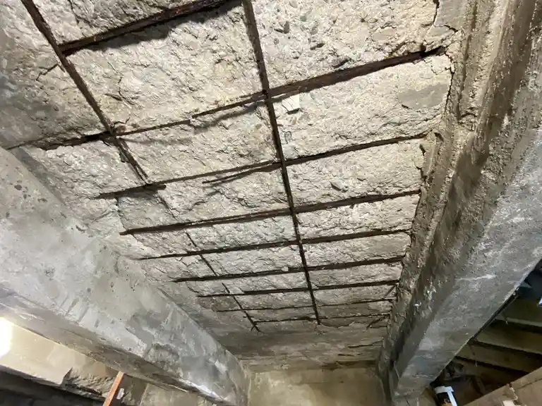 A close-up, upward-looking shot of a rough, unfinished concrete ceiling with visible rusted rebar forming a grid pattern…