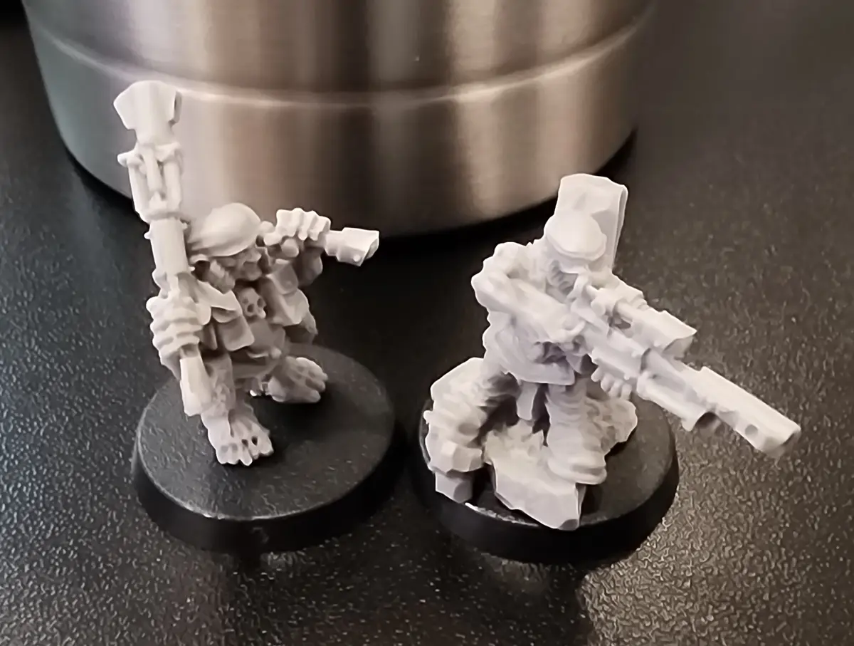 Two unpainted white miniature figurines of soldiers with weapons stand on black circular bases on a dark speckled surface…