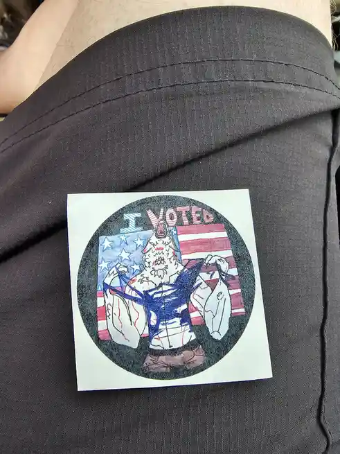 A hand-drawn 'I VOTED' sticker is stuck on a person's black shirt. The sticker features a muscular, bearded, wolf-like…