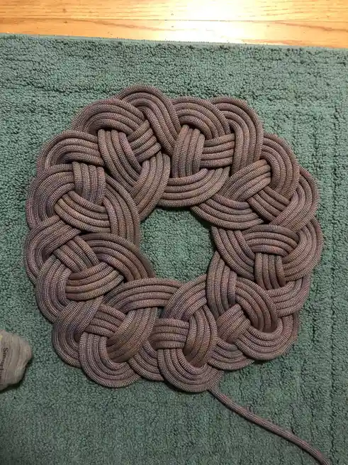 A circular, intricately woven rope wreath in brown and purple hues rests on a green shag rug, with a wooden floor visible at…