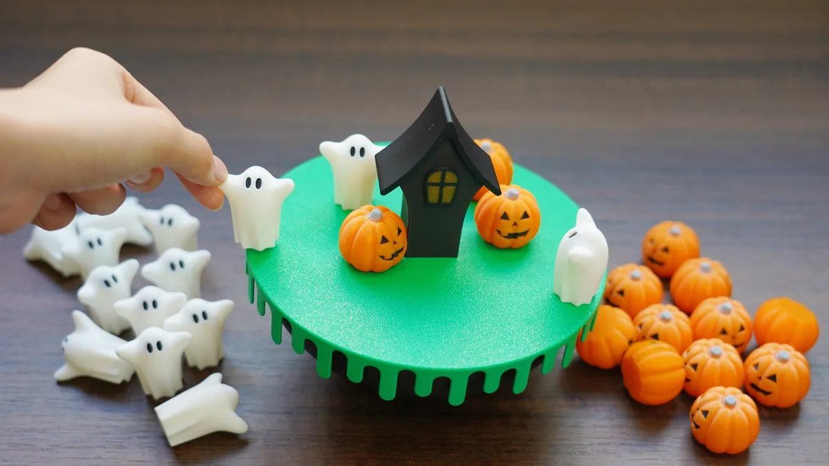 A hand places a small white ghost figurine onto a green circular base that resembles a haunted house scene with a black…