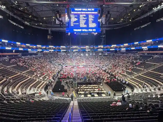 A wide-angle photo of a large, mostly empty arena with a few scattered people, featuring a massive central scoreboard…