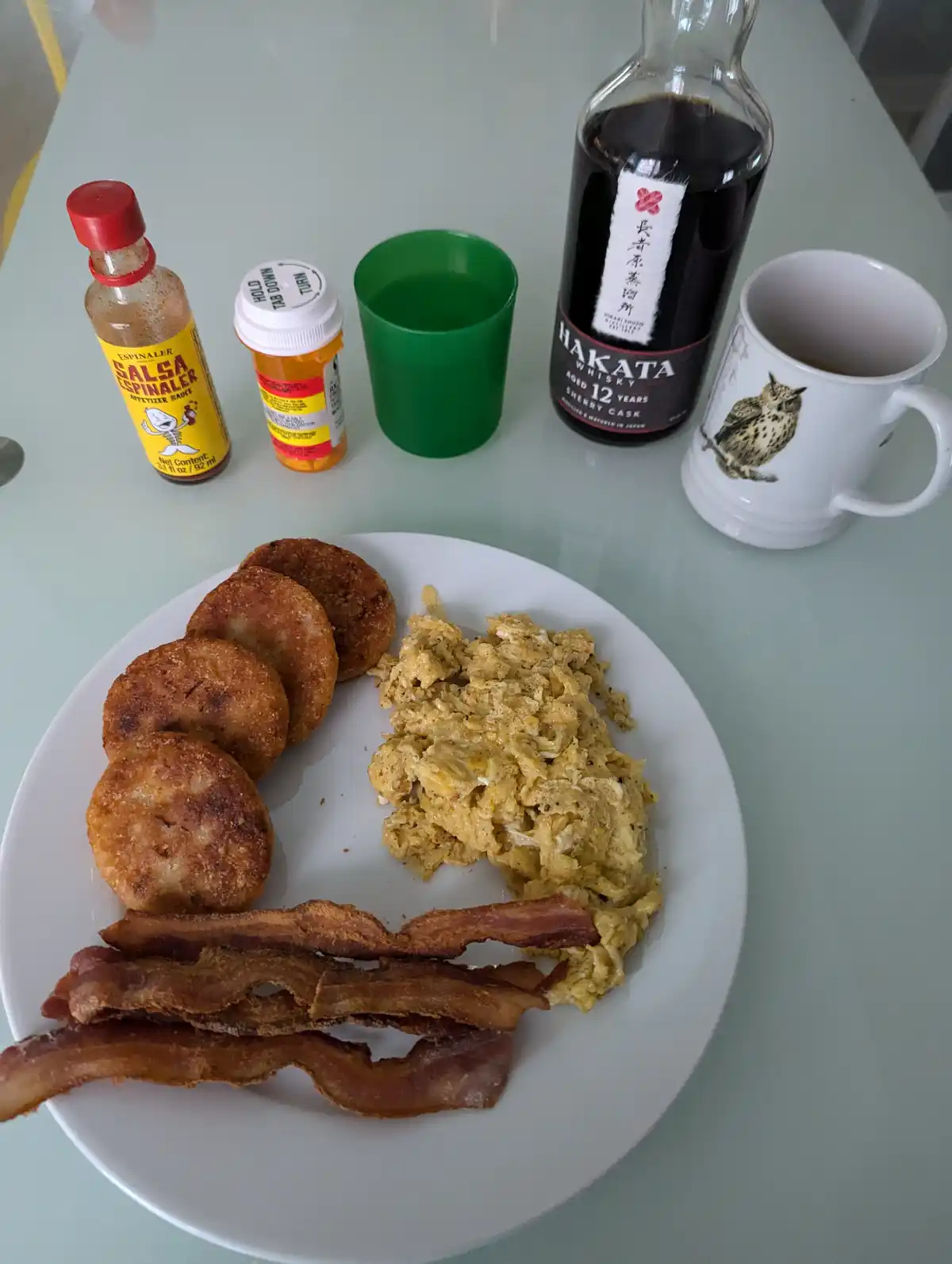A plate of breakfast food including scrambled eggs, bacon, and fried potato patties sits on a table next to a bottle of…