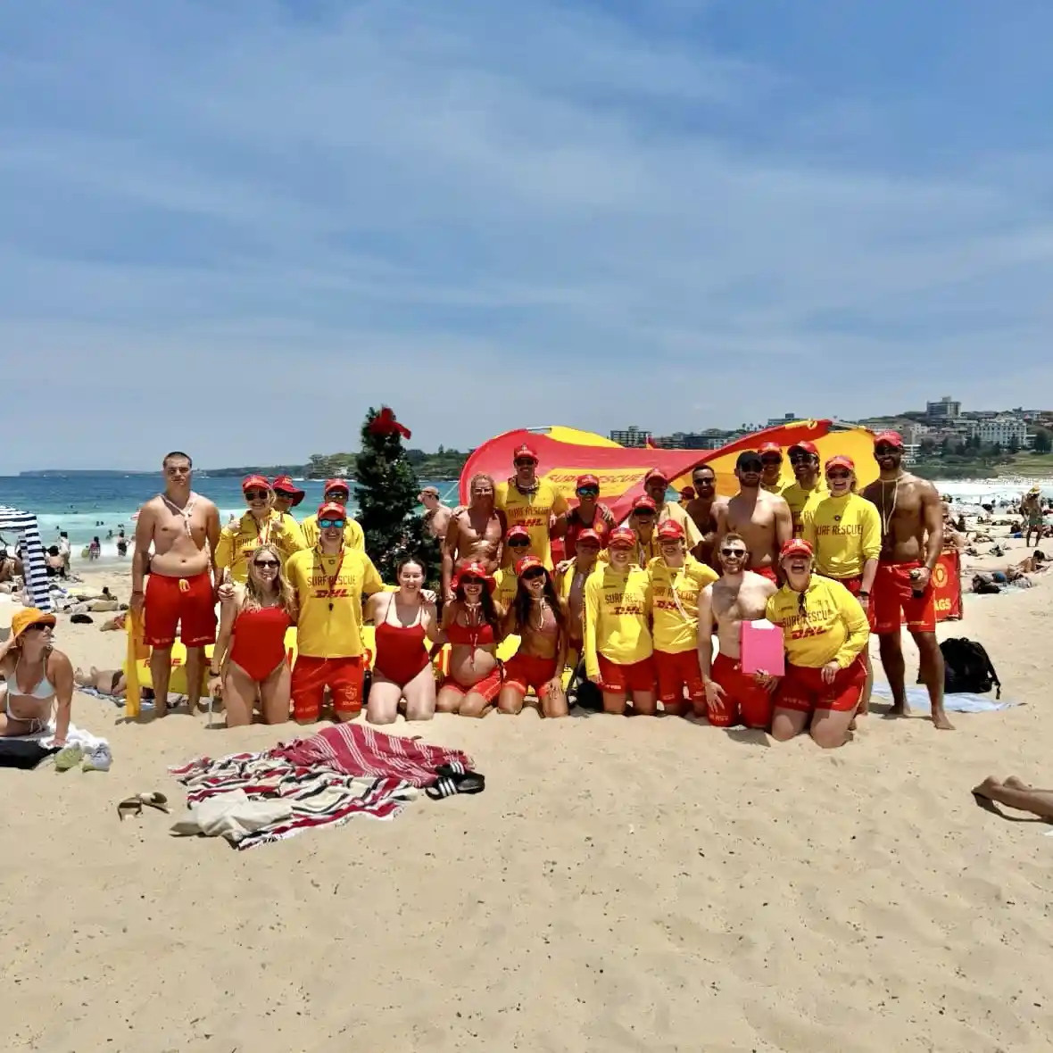 A group of surf lifesavers in yellow shirts and red shorts pose for a beach photo, with a small decorated christmas tree…