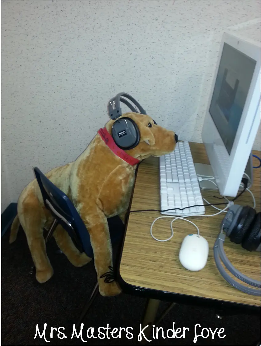 A tan stuffed dog sits upright on a blue school chair at a wooden desk, wearing gray over-ear headphones and a red collar…