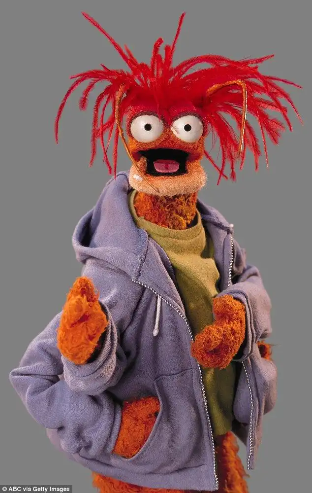 An orange Muppet with wild red hair and a surprised expression wears a purple hoodie over a green shirt, hands in pockets…