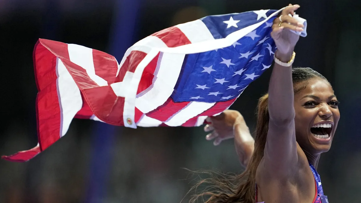 A jubilant female athlete, likely from the USA, is captured mid-celebration, holding a large American flag aloft with one…