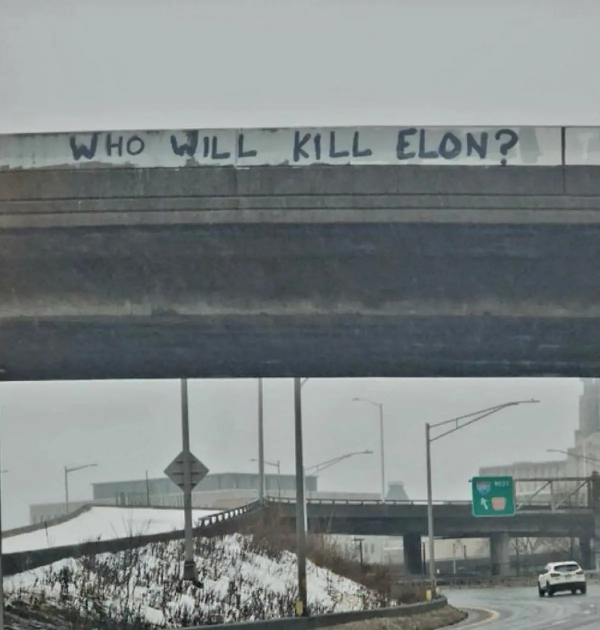 A photograph of a snowy highway underpass with graffiti painted in black on the concrete overpass reading 'WHO WILL KILL…