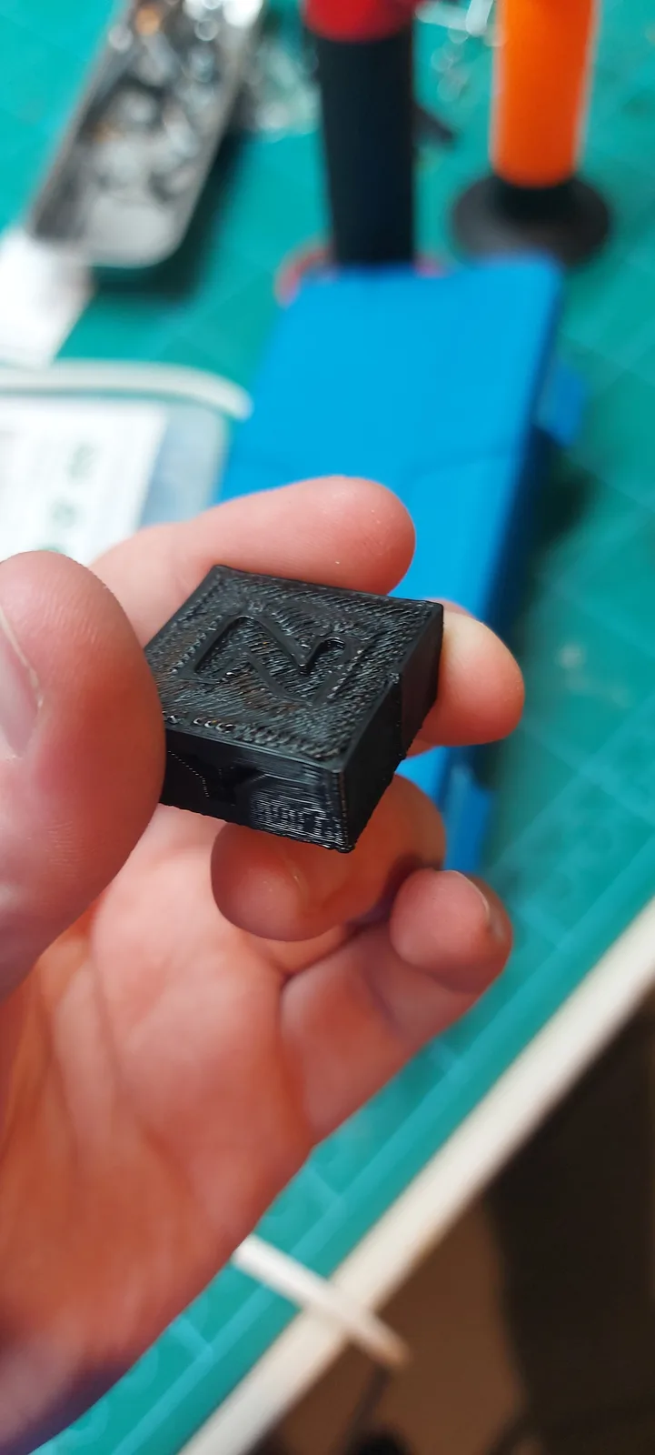 A hand holds a small, black, 3d-printed cube with the letter 'N' embossed on its top face. The object has visible layer…