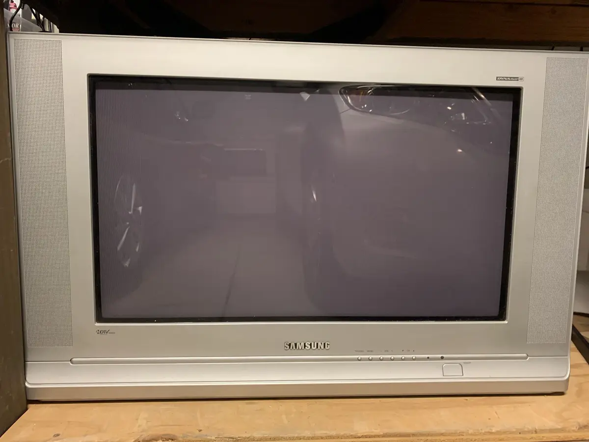A silver Samsung CRT television sits on a wooden surface, its screen off and reflecting a car and surroundings. The overlaid…