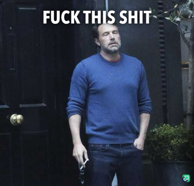 A man in a blue sweater and jeans stands in front of a dark door, looking exhausted and annoyed, holding a cigarette. The…