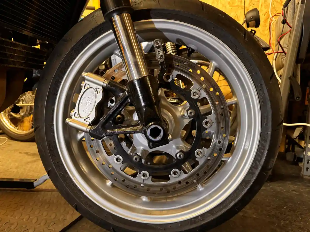 A close-up photo of a motorcycle's front wheel showing a newly installed brake caliper and rotor, with the brand 'HEL'…