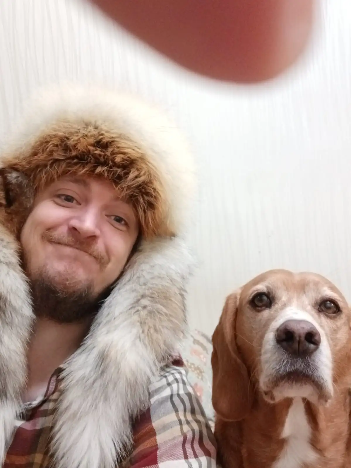 A man with a beard wearing a furry winter hat and coat takes a selfie with a beagle dog beside him. The man has a smug…