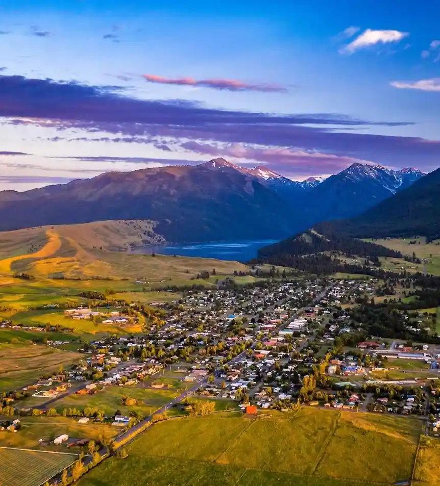 An aerial photograph shows a small town nestled in a valley surrounded by rolling green hills, a large lake, and snow-capped…