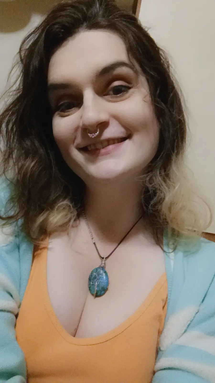 A smiling woman with dark curly hair and a nose ring wears an orange top under a light blue cardigan. She has a large, oval…
