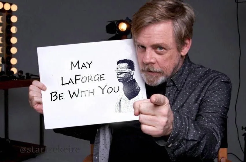 A man with gray hair and a beard, wearing a dark patterned shirt, holds up a white sign that reads 'May LaForge Be With You'…