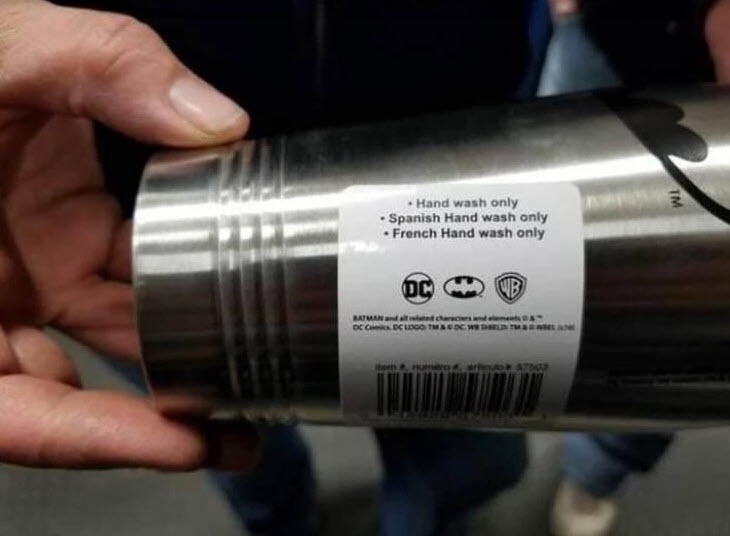 A close-up of a person holding a silver Batman-themed thermos, showing its label which lists absurd washing instructions…