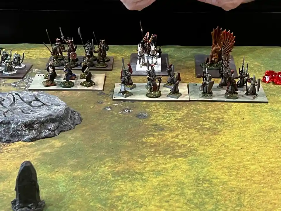 A tabletop wargaming scene shows multiple armies of painted miniatures arranged in battle formations on a textured green and…