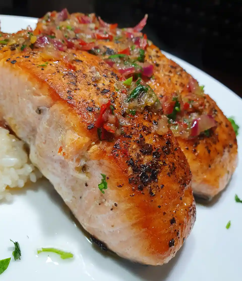 A close-up photo of a perfectly seared salmon fillet on a white plate, topped with a colorful salsa of chopped red onions…
