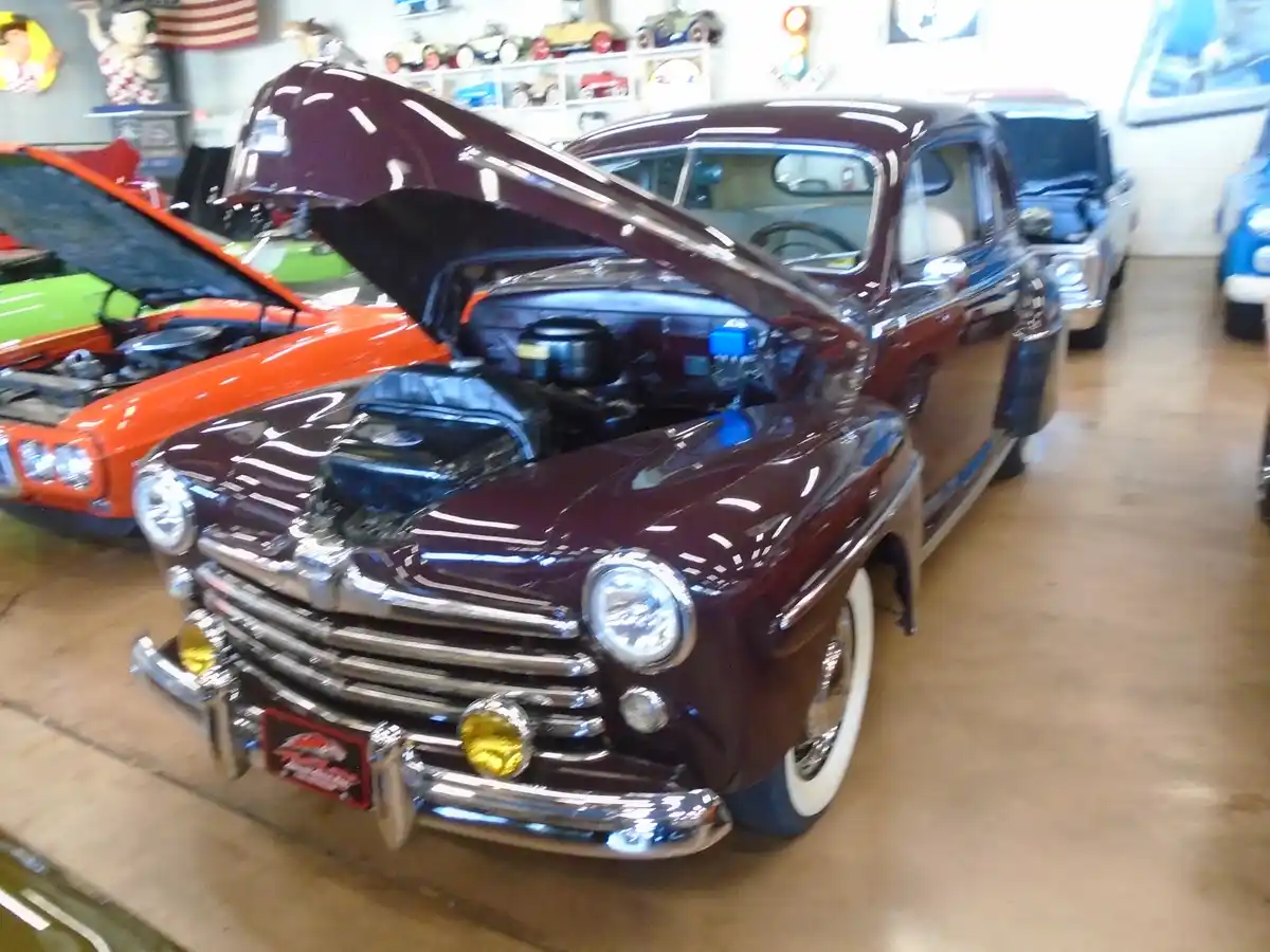 A maroon classic car with its hood open is displayed in a garage alongside other vintage vehicles, including an orange car…