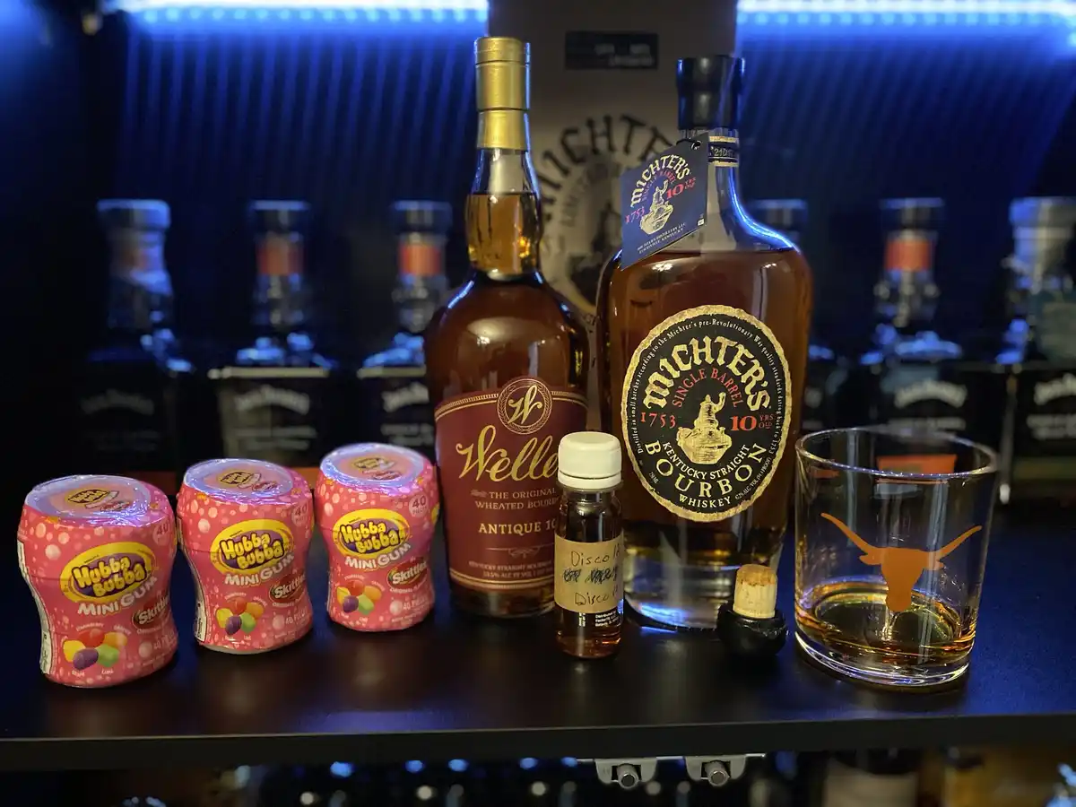 A bar shelf displays three pink containers of Hubba Bubba Mini Gum Skittles, two bottles of bourbon (Weller Antique 10 and…