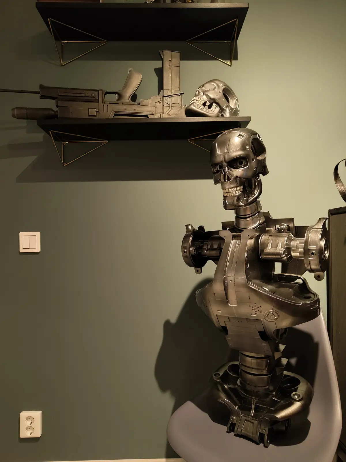 A metallic Terminator-style robot bust sits on a modern chair, with shelves above holding a replica gun and a skull. The…