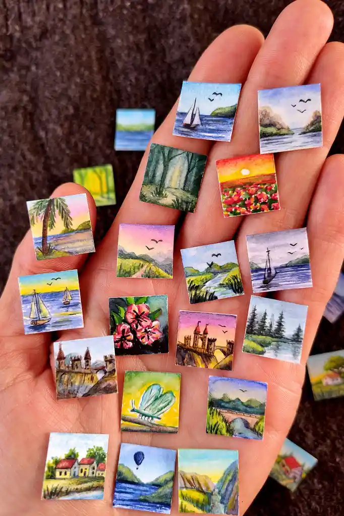 A hand holds numerous tiny square watercolor paintings depicting various scenic landscapes like sailboats, forests, sunsets…