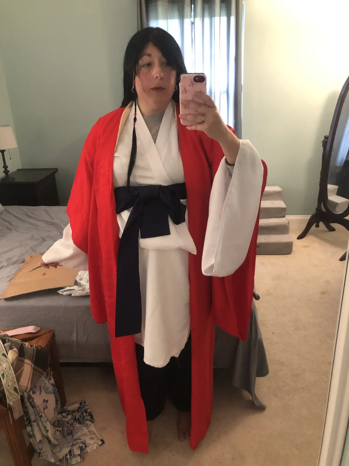 A person with dark hair takes a mirror selfie in a bedroom while wearing a red and white kimono-style outfit with a navy…