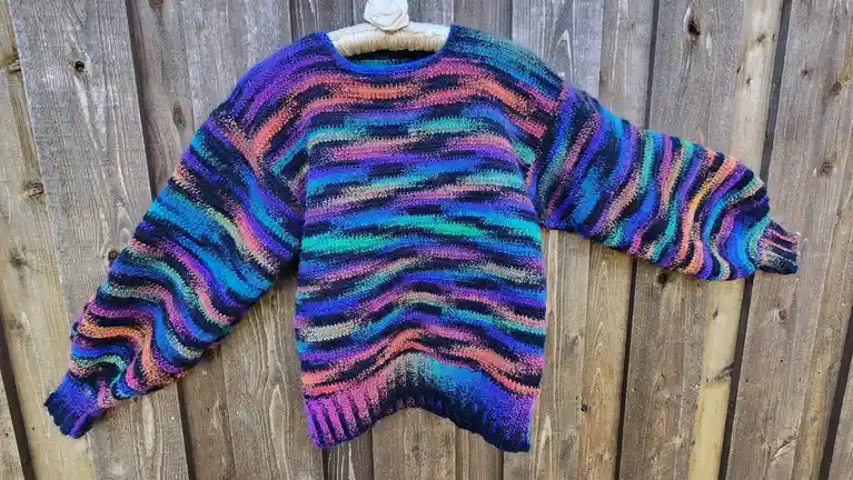 A hand-knitted, multicolored sweater with wavy horizontal stripes of purple, blue, teal, pink, and orange hangs on a wooden…
