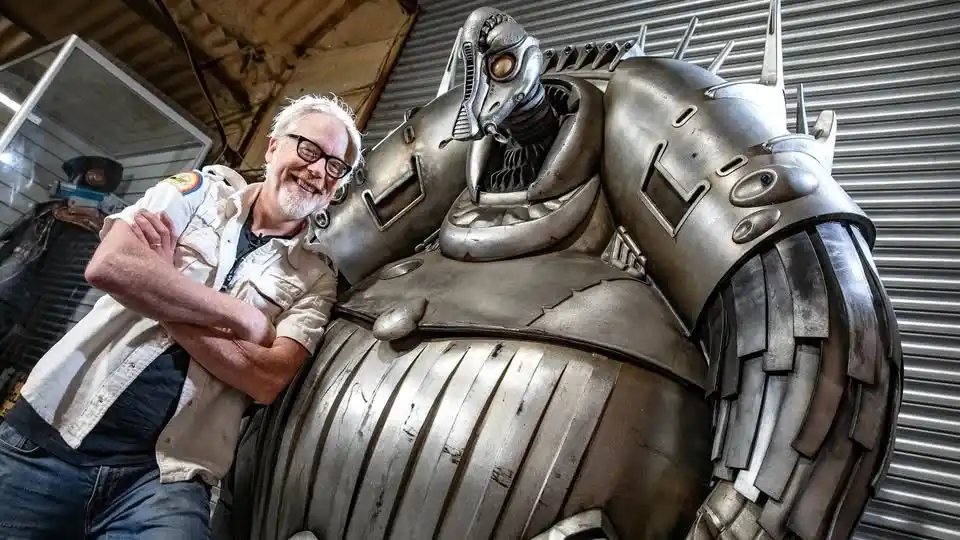 A smiling older man with glasses and a beard stands confidently with arms crossed beside a massive, detailed, metallic robot…