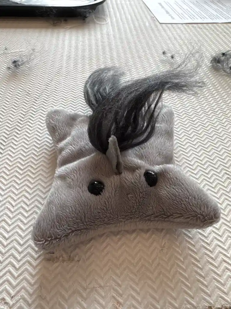 A gray plush unicorn head with a messy black wig is lying on a textured white surface, surrounded by stray fibers. The text…