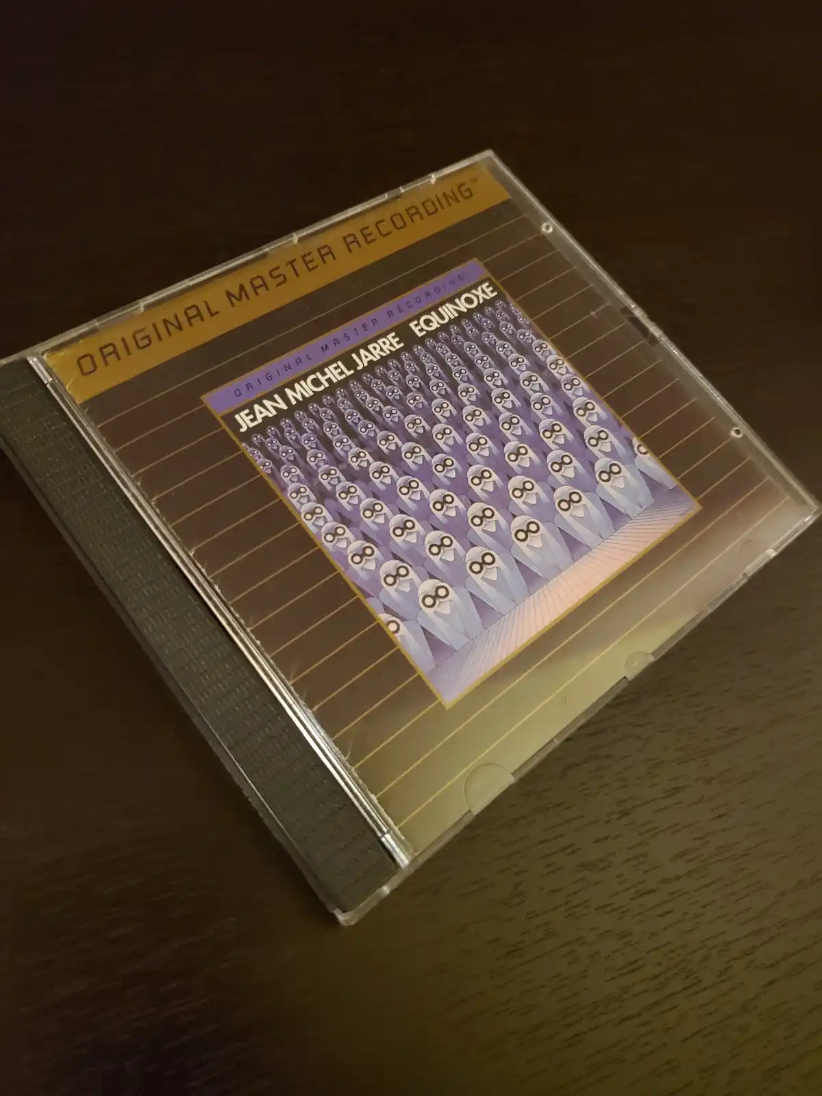 A physical CD case for Jean Michel Jarre's album 'Equinoxe' rests on a wooden surface. The cover art features a repeating…